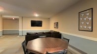 This single-floor space is designed as a versatile recreational and entertainment area, offering a welcoming atmosphere suited for gatherings and leisure. The room features neutral-toned walls with a two-tone finish, separated by a clean horizontal chair rail detail that adds a subtle architectural touch to the interior.

At the heart of the space sits a large octagonal game table surrounded by dark wood chairs, making it an ideal setting for card games, board games, or casual dining. Behind it, a dark leather sectional sofa provides generous seating and is positioned to face a wall-mounted flat-screen television, creating a dedicated media viewing area. A framed photo collage on the adjacent wall adds a personal, decorative element to the room.

The space is finished with smooth, light-colored flooring that complements the warm tones of the walls, and recessed ceiling fixtures provide consistent, even illumination throughout. The layout is open and functional, allowing the room to serve dual purposes as both a media lounge and a game room. Overall, the space conveys a relaxed, casual ambiance well-suited for entertainment and social activity.