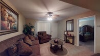 This single-floor residence offers a well-organized layout with distinct living, dining, sleeping, and cooking areas. The living room serves as the central gathering space, featuring carpet flooring, a ceiling fan with light fixture, and vertical blinds that filter natural light. Dark upholstered sofas and a glass-top coffee table furnish the room, complemented by framed artwork and indoor greenery. A dedicated dining area accommodates a round glass-top table with traditional wooden chairs beneath a flush ceiling light, with chair rail molding adding a classic architectural detail. The galley-style kitchen is lined with wooden cabinetry on both sides, offering ample storage, laminate countertops, a double basin sink, electric coil range, refrigerator, and a dishwasher. Two carpeted bedrooms provide quiet retreats, each with windows fitted with blinds for privacy. The bathroom includes a wooden vanity with granite-style countertop, a large mirror with overhead lighting, tile flooring, and a full-size tub and shower combination with white tile surround. Throughout, the warm neutral tones and traditional furnishings contribute to a cohesive, understated aesthetic.