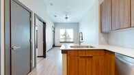 This single-floor unit showcases a contemporary open-concept design that seamlessly connects the kitchen and living areas. The kitchen serves as a central hub, anchored by a substantial island with a white quartz countertop, an undermount sink, and a pull-down faucet. Warm wood cabinetry lines the upper and lower walls, complementing the stainless steel appliances, including a refrigerator, range, and over-the-range microwave. A subway tile backsplash adds a clean, classic touch.

Light wood-look vinyl flooring flows throughout the main living spaces, enhancing the bright, airy atmosphere created by white walls and recessed lighting. A ceiling fan in the living area adds comfort and functionality. Dark-framed interior doors provide a striking contrast against the neutral palette, lending a modern edge to the design.

The bathroom features matching wood cabinetry with white countertops, a soaking tub with tiled surround, and direct access to a dedicated walk-in closet outfitted with built-in shelving and a closet organizer system. The carpeted bedroom offers a comfortable retreat, highlighted by a set of full-length double doors with transom windows above, allowing natural light and outdoor views to fill the room.