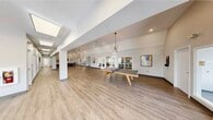 This generously sized single-floor amenity facility spans an impressive footprint, offering a diverse range of spaces designed to foster community, wellness, and recreation. The centerpiece of the facility is a vast, open-concept recreation and event hall featuring light wood-look flooring, soaring vaulted ceilings, and statement sputnik chandelier fixtures that lend a contemporary flair. Skylights and recessed lighting ensure the space remains bright and welcoming throughout. A ping-pong table and ample open floor area make this hall ideal for social gatherings and activities. Adjacent to the main hall is a comfortable lounge area furnished with a sectional sofa, wall-mounted TV, and framed artwork, creating an inviting retreat. The full kitchen is outfitted with stainless steel appliances, granite countertops, warm wood cabinetry, and generous natural light from large windows. The fitness center is well-appointed with a comprehensive array of cardio machines and strength training equipment on durable rubber flooring, illuminated by large skylight panels. Additional smaller rooms offer intimate round-table meeting spaces, each tastefully decorated with curated artwork. Throughout, the design is clean, modern, and cohesive, with neutral tones, wood-look flooring, and contemporary fixtures creating a unified and polished atmosphere.