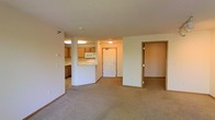 This single-floor unit offers a practical and functional layout centered around an open-concept living and kitchen area. The spacious carpeted living room provides a comfortable and neutral foundation, with warm beige tones throughout that create a cohesive, understated ambiance. A kitchen peninsula with a light-colored countertop separates the kitchen from the living space, offering both a workspace and a casual dining or prep area. The kitchen is equipped with wood-toned cabinetry and includes a built-in microwave, with tile flooring that contrasts neatly with the carpeting in the living area. A textured ceiling runs throughout the unit, adding subtle visual depth. The front entry door features a classic paneled design and is centrally visible from the living room, contributing to the open sightlines of the space. Two additional doorways are accessible from the main living area, leading to further rooms within the unit. Flush-mounted ceiling light fixtures provide consistent overhead lighting throughout. Overall, the space presents a clean, move-in-ready environment with a simple and functional design.