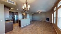This two-floor space is designed with an open-concept layout that seamlessly connects the kitchen, dining, and living areas, creating a spacious and functional flow throughout. The kitchen is appointed with dark wood cabinetry, stainless steel refrigerator, and a peninsula island topped with granite-style countertops — ideal for both meal preparation and casual dining. A statement wrought-iron chandelier with frosted glass shades anchors the dining area, adding a touch of elegance to the space. The living area is generously sized, bathed in a neutral, warm color palette with soft gray walls that complement the earthy tones of the large-format tile flooring found throughout the main level. A ceiling fan overhead ensures comfortable airflow, while a sleek built-in fireplace on the far wall serves as a refined focal point for the living space. Recessed lighting and ample ceiling vents contribute to both the aesthetic and the practicality of the environment. The open sightlines between rooms enhance the sense of scale, making the space feel bright and welcoming. Overall, this property offers a clean, modern aesthetic with functional design elements that cater to comfortable everyday living across two floors.