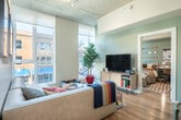 Located at 301 N Broom St in downtown Madison, WI, this six-bedroom, three-bathroom unit offers a well-designed layout across a single floor. The open-concept kitchen and living area serves as the heart of the space, featuring light wood cabinetry, white quartz countertops, stainless steel appliances, a breakfast bar with seating, exposed concrete ceiling details, and pendant lighting. The adjacent living room is bathed in natural light through floor-to-ceiling windows that frame views of the surrounding urban streetscape. Light wood flooring runs throughout, lending warmth and continuity to the space. The six bedrooms are comfortably proportioned, each with access to natural light via large windows. Three hallways connect the private rooms efficiently throughout the unit. The three bathrooms are clean and contemporary, with one featuring a glass-enclosed walk-in shower with built-in shelving and light wood vanity accents. Additional hallway storage with light wood cabinetry adds practical functionality. The overall aesthetic is modern and cohesive, with a neutral palette, clean lines, and an abundance of natural light, making this a well-suited space for shared living in the heart of Madison.