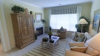 This single-story living space exudes a cozy, traditional charm with a carefully curated country aesthetic. The room is anchored by a classic wood-burning fireplace with a white tile surround and a simple mantel adorned with candles and decorative greenery. Flanking the fireplace is a substantial pine armoire with raised panel doors and lower drawers, providing ample storage while adding rustic warmth to the space.

A large picture window dressed with a blue-and-white plaid Roman shade and white blinds allows generous natural light to fill the room, illuminating the soft yellow walls accented by white crown molding. The seating area features a wicker-style armchair with plush cushions and throw pillows, complemented by an upholstered sofa, both arranged around a rectangular coffee table styled with decorative boxes and a fruit centerpiece.

A textured striped area rug grounds the seating arrangement, while potted plants and floral accents throughout the space add life and color. The overall ambiance is inviting and lived-in, blending functional storage with decorative touches for a comfortable, homey atmosphere.