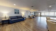 This generously sized single-floor community space is designed with a relaxed coastal aesthetic, evident in its soft blue-gray walls, nautical artwork, and light hardwood-style flooring that extends throughout the entire area. The open layout is both versatile and welcoming, accommodating multiple functional zones within one expansive room.

The lounge area is furnished with navy blue sofas and accent chairs arranged around coffee tables, with a wall-mounted flat-screen television and decorative pieces including a compass rose wall accent and sailboat artwork. Recessed lighting and table lamps provide warm, layered illumination throughout.

Along one side of the room, a bar-height counter with tall wooden stools offers an additional gathering spot, backed by a decorative tiled accent wall in coastal tones. A small service area with cabinetry is accessible behind the counter. Large windows fitted with horizontal blinds allow natural light to fill the space, and a tall potted plant adds a touch of greenery.

The entrance features double glass-panel doors that enhance the sense of openness and allow further natural light inside. The textured ceiling with recessed lighting adds dimension overhead. Overall, the space presents a clean, comfortable, and functional environment suited for community gatherings and social events.