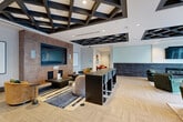 Located at 60 11th St NE in Atlanta's vibrant Midtown neighborhood, this upscale community lounge offers a thoughtfully designed single-floor layout that balances social and relaxed environments. The space is anchored by a striking kitchen bar area featuring an expansive marble-topped island with stylish leather and wood barstools with gold-accented legs, glass-front cabinetry, a tile backsplash, a wine refrigerator, and a wall-mounted TV — ideal for gatherings and entertaining.

The main living area showcases a bold architectural coffered ceiling with dark geometric lattice detailing, creating visual depth overhead. A wood-paneled accent wall frames a sleek linear fireplace beneath a large wall-mounted TV, while a plush leather sofa, curated armchairs, and a patterned area rug complete the lounge atmosphere. Floor-to-ceiling windows invite abundant natural light and offer views of the adjacent outdoor terrace and Atlanta's cityscape.

A secondary lounge area provides a quieter setting with upholstered seating, built-in workstations, and vibrant abstract artwork along the walls. Large glass doors open directly to a furnished outdoor patio. Throughout, light herringbone-patterned flooring ties the spaces together, lending a cohesive, contemporary elegance to this well-appointed amenity space.