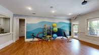 Located at 1000 Flycatcher Way in Arden, NC, this fitness center offers a thoughtfully designed single-floor space dedicated to health and wellness. The room is immediately distinguished by a sweeping, floor-to-ceiling mural depicting the iconic Blue Ridge Mountains in layered shades of blue — a nod to the surrounding Western North Carolina landscape that lends the space both energy and character.

Warm hardwood flooring extends throughout, providing a clean, durable surface suitable for a wide range of activities. Recessed lighting and a ceiling fan with a sleek black fixture ensure the space remains bright and comfortable during workouts. Crown molding and crisp white trim add a polished, refined aesthetic.

A full-wall mirror along one side of the room enhances the sense of openness and supports functional training and form monitoring. Large windows and a glass-paned exterior door invite abundant natural light and offer views of the surrounding greenery, creating an airy atmosphere.

The space is equipped with stationary cycling machines, an adjustable workout bench, a wall-mounted flat-screen television, and a well-stocked rack featuring resistance equipment, exercise balls, dumbbells, and other fitness accessories. A balance trainer and additional freestanding equipment round out the offerings. This fitness center provides residents with a comprehensive and inviting environment to support an active lifestyle.