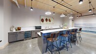 Located at 310 N Reid Pl in Sioux Falls, SD, this single-floor space offers a thoughtfully designed open-plan layout that seamlessly integrates a kitchen and living area. The interior is defined by a striking combination of natural wood and industrial elements — warm, light-toned wood plank ceilings contrast beautifully with polished concrete floors, creating a refined yet approachable aesthetic throughout.

Structural wood beams and columns add architectural character to the open layout, while exposed piping along the ceiling reinforces the industrial undertone. Pendant lighting in varied styles is suspended throughout, providing layered illumination across the space.

The kitchen features sleek gray cabinetry with white countertops, a spacious central island with bar seating for several guests, a beverage refrigerator, and ample counter space for preparation and entertaining. A large-scale botanical artwork anchors the kitchen wall, adding a warm, curated touch.

The living area is anchored by a full-wall scenic mural rendered in soft blue and gray tones, creating a distinctive focal point alongside a wall-mounted flat-screen television. Comfortable seating arrangements, a woven coffee table, and patterned accent chairs contribute to a relaxed, welcoming atmosphere. Large windows and a glass door invite natural light and provide views of the exterior surroundings, connecting the interior to the outdoors.