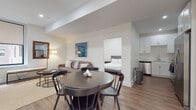 This single-floor residence features a thoughtfully designed open-concept layout that seamlessly connects the living, dining, and kitchen areas. The living room is anchored by a tufted brown sofa accented with layered throw pillows and a woven area rug, complemented by a round wood coffee table and contemporary floor lamp. A large framed blue artwork adds a bold focal point to the space. A window with roller shades allows natural light to filter in while offering views of the surrounding urban streetscape.

The dining area features a round dark wood table surrounded by classic spindle-back chairs, flowing naturally into the kitchen. The kitchen is fitted with white shaker-style cabinetry, light countertops, and stainless steel appliances including an electric range, over-the-range microwave, and dishwasher.

The unit includes two bedrooms, each finished with light hardwood flooring, neutral white walls, and tufted gray upholstered bed frames paired with crisp white bedding. One bedroom features a sliding frosted-glass closet door, while the other benefits from a large window with urban views.

The bathroom is finished with marble-style porcelain tile flooring, white subway tile surrounding the soaking tub and shower combination, and a white vanity with chrome fixtures, creating a clean, polished aesthetic throughout.