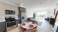 Located at 14528 Erwin St in Los Angeles, CA, this single-floor unit offers a thoughtfully designed open-concept layout finished with light hardwood flooring and recessed lighting throughout. The kitchen is a standout feature, outfitted with two-tone cabinetry in slate gray and light taupe, white quartz countertops, a stainless steel undermount sink, and a full suite of stainless steel appliances including a gas range, built-in microwave, dishwasher, and French door refrigerator. The kitchen flows seamlessly into a dedicated dining area and an adjacent living room, which is bathed in natural light from large sliding glass doors that open to an outdoor balcony with open city views. The bedroom is bright and airy, featuring a sizable window framing elevated views of the surrounding Los Angeles skyline, complemented by recessed lighting and ample floor space. A frosted glass door leads from the bedroom to the well-appointed bathroom, which includes a white vanity with dark countertop, an illuminated mirror, and a tub-shower combination with glass enclosure and tiled surround. The overall ambiance is clean, contemporary, and light-filled.