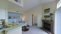 This single-story space is defined by its impressive vaulted ceilings, which lend an airy, open quality to the living area. The main living room is carpeted and anchored by a classic brick fireplace with a traditional mantel, serving as a natural focal point. A built-in ledge or half-wall separates the living area from what appears to be a kitchen or entry zone, enhancing the open-concept flow while maintaining subtle spatial definition. Neutral, warm-toned walls complement the light carpeting throughout, creating a cohesive and welcoming atmosphere. A doorway visible from the main living space leads to additional rooms, suggesting a well-connected interior layout. Ceiling vents are integrated throughout, indicating a central HVAC system. Wall-mounted décor niches and accent lighting add character to the space. The combination of high ceilings, soft neutral tones, and the brick fireplace gives the living area a comfortable yet spacious ambiance suited for everyday living and entertaining.