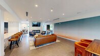Located at 10 Patriot Pkwy in Weymouth, Massachusetts, this single-floor amenity space offers a generously sized, open-plan layout that seamlessly integrates a living lounge and a fully equipped kitchen. The living area is thoughtfully designed with a striking ombre accent wall that transitions from deep teal to warm peach, creating a vibrant yet relaxed atmosphere. A linear fireplace anchors one wall beneath a mounted flat-screen television, surrounded by comfortable seating including leather sofas, navy armchairs, and woven ottomans arranged over a colorful area rug atop large-format hexagonal tile flooring. A billiards table adds a recreational element to the space. The kitchen features sleek, slate-blue cabinetry, light quartz countertops, a bold teal penny-tile backsplash, and stainless steel appliances including a gas cooktop with overhead range hood and built-in wall ovens. A cozy dining nook with banquette seating and pendant lighting adjoins the kitchen, while a large window offers views of the surrounding greenery. Recessed lighting throughout and a linear HVAC system maintain a clean, modern ceiling profile. The space is well-suited for gathering, entertaining, and everyday community use.