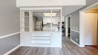 Located at 1300 Nashville Pike in Gallatin, Tennessee, this well-appointed one-bedroom, one-bathroom unit offers a thoughtfully designed single-floor layout. Wood-look flooring flows consistently throughout the space, complemented by a neutral gray color palette and crisp white trim, including a distinctive chair rail detail visible in several rooms.

The kitchen is a standout feature, positioned behind a striking white-framed open partition wall with built-in shelving — a charming architectural detail that connects the kitchen to the living area while maintaining definition. The kitchen itself is equipped with white cabinetry, light countertops, a stainless steel sink, stainless appliances including a range and microwave, and a refrigerator. A skylight-style ceiling fixture adds brightness overhead.

The bedroom is a comfortable size, featuring a ceiling fan with light fixture, a window that brings in natural light, and the same consistent flooring and gray tones found throughout the unit. The bathroom includes a full tub/shower combo, a vanity with storage, and wood-look flooring.

A dedicated laundry room with a full-size washer and dryer provides added everyday convenience. A hallway connects the rooms efficiently, completing this compact yet fully functional layout.