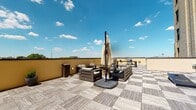 Located in the heart of Richmond, Virginia, this rooftop terrace at 213 E Broad St offers an exceptional outdoor experience in a prime urban setting. The space is finished with a stylish interlocking wood-tile deck that adds warmth and texture underfoot, creating a polished yet relaxed atmosphere open to the sky.

The terrace is thoughtfully arranged for both lounging and entertaining. A comfortable seating area features dark wood-framed chairs and a sofa with cushioned upholstery, centered around a low coffee table and complemented by a large canopy umbrella — ideal for shaded relaxation on sunny days. A dedicated grilling station positioned along the perimeter makes the space well-suited for outdoor dining and social gatherings.

Perimeter walls in a warm ochre tone provide a sense of enclosure while preserving the open, airy feel of the rooftop. Expansive, unobstructed views stretch out in multiple directions, offering broad sightlines of the surrounding Richmond cityscape and beyond. The combination of natural light, open sky, and thoughtful furnishings gives the terrace a versatile ambiance — equally suited for casual afternoon lounging or lively evening entertaining. This rooftop terrace stands as a distinctive outdoor amenity in one of Richmond's most accessible and vibrant downtown locations.