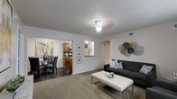 This single-floor space showcases an open-concept design that seamlessly connects the living room, dining area, and kitchen into one cohesive and functional layout. The living room is anchored by plush carpeting, a sleek dark sofa, and a contemporary white coffee table, complemented by decorative metallic wall art that adds a modern touch. A ceiling fan with a light fixture provides both comfort and illumination.

The dining area, visible to the left, features dark hardwood-style flooring and a glass-top dining table paired with upholstered chairs, creating an elegant contrast to the living space. A framed artwork piece in warm yellows and whites adds a vibrant accent to the dining nook.

The kitchen, accessed through a wide pass-through opening, is outfitted with warm wood cabinetry, a stainless steel microwave, and a breakfast bar that enhances the open flow between spaces. A textured popcorn ceiling runs throughout, and neutral white walls allow the furnishings and décor to take center stage. The overall ambiance is modern, clean, and welcoming, making efficient use of the available space.