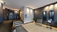 The lobby at 5160 Avenue Gatineau in Montréal, Québec, serves as the welcoming point of entry for this residential building. The space is organized into a two-stage entry sequence, beginning with a secure glass-and-metal vestibule equipped with an intercom and access control panel set against painted blue brick walls. Large glass doors flood this entry zone with natural light, offering views of the surrounding streetscape.

Stepping inside, the main lobby area features light-toned large-format tile flooring that transitions to carpeted surfaces toward the interior staircase. The walls are finished in a contemporary combination of deep charcoal grey and crisp white, accented by painted blue-grey brick detailing that lends the space an urban, textured character. Track lighting mounted to the ceiling provides focused illumination throughout.

A modest seating area with a tufted grey chair, a potted plant, and a full-length mirror adds a welcoming touch to the interior lobby. A staircase with dark metal railings connects the lobby level to the building's hallway corridor, which is lined with carpeting, painted brick walls, and sconce lighting leading to individual unit doors. Security and building information panels are conveniently positioned near the inner entrance. Overall, the lobby presents a functional yet thoughtfully styled space that reflects a well-kept residential building.