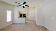 This single-floor residence is designed with an open, airy layout that maximizes the sense of space throughout. The living area immediately draws the eye upward to impressively high ceilings, which are complemented by a large ceiling fan that enhances airflow and comfort. Neutral-toned carpeting extends across the floor, lending a warm and cohesive feel to the main living space.

The kitchen is thoughtfully integrated into the open floor plan, separated from the living area by a raised breakfast bar that provides both a casual dining option and a natural visual boundary. Light wood cabinetry with recessed lighting overhead adds warmth to the kitchen, while black appliances — including a full-size refrigerator and microwave — offer a sleek contrast. The layout ensures the kitchen remains accessible and sociable without being fully exposed to the main living area.

A pendant chandelier hangs elegantly between the kitchen and what appears to be a dining or entry area, adding a refined decorative touch. The overall color palette is soft and neutral, with cream-toned walls throughout that reflect light and contribute to the bright, welcoming ambiance. The single-floor layout ensures easy, step-free navigation throughout the entire space, making it both functional and inviting.