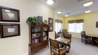 This single-floor common room offers a warm and inviting atmosphere designed for relaxation and recreation. The space is anchored by a richly stained wooden bookcase with glass-paneled doors, displaying an organized collection of books and decorative items, including a lush potted fern that adds a touch of greenery. Framed artwork depicting playing card suits adorns the soft yellow walls, complementing the room's game-centric theme.

At the center of the room sits a classic wooden game table topped with a checkerboard surface, surrounded by cushioned leather chairs with nailhead trim detailing, lending an air of traditional elegance. Natural light filters generously through multiple windows dressed with fabric valances in warm, patterned tones, brightening the neutral carpeted floor throughout.

Flush-mount ceiling fixtures provide consistent ambient lighting, ensuring the space remains well-lit during evening hours. A cozy seating nook positioned near the windows offers an additional spot for quiet reading or conversation. The overall ambiance is comfortable and homey, thoughtfully furnished to encourage social interaction and leisurely activities.