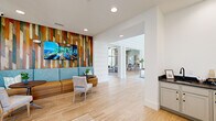 This single-floor lobby and lounge space exudes a refined coastal aesthetic, combining architectural detail with thoughtful interior design. Soaring ceilings clad in white shiplap paneling create a dramatic sense of vertical scale, while light wood flooring runs throughout, lending warmth and continuity to the open layout. A striking suspended canoe pendant fixture serves as a focal point above the central corridor, reinforcing the nautical theme carried across the space.

Nautical-themed artwork, including a vibrant canoe painting and framed fishing imagery, adorns the crisp white walls, complementing the coastal character of the design. Large windows and glazed doors invite abundant natural light, further enhancing the airy, welcoming atmosphere.

A dedicated seating lounge features a bold, multicolored reclaimed wood accent wall paired with a wall-mounted display, flanked by a contemporary blue and tan banquette with coordinating side chairs and tables — ideal for casual gatherings or quiet conversation. An adjacent sitting area offers mid-century modern accent chairs, soft textiles, and a compact refreshment station, providing a comfortable retreat.

The space connects seamlessly to additional amenity areas visible through open archways, including a lounge and dining area, offering a cohesive flow throughout. The overall ambiance is clean, bright, and inviting, with a consistent coastal-resort character throughout.