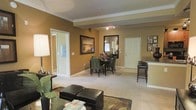 This single-floor residence features an open-concept layout that seamlessly connects the living and dining areas, creating a cohesive and inviting atmosphere. The warm golden-tan walls are complemented by crisp white crown molding, lending the space a polished, classic character throughout.

The living area is anchored by plush carpeting in a neutral tone, providing a soft foundation for the dark leather ottoman seating and a patterned area rug. A side table with decorative accents and greenery adds a touch of warmth to the corner. The space transitions naturally into the dining area, where a dark wood table is surrounded by upholstered green chairs beneath a ceiling-mounted spotlight track, offering focused ambient lighting.

A large framed landscape artwork adorns the dining wall, while a tall rectangular mirror enhances the sense of depth and reflects natural light into the space. A counter-height bar area with dark stools is visible to the right, suggesting a kitchen pass-through or breakfast bar nearby.

Doorways lead to additional rooms, including what appears to be a well-lit bathroom or hallway, and a private room with a white paneled door, reinforcing the practical and comfortable nature of this residence.