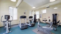 This fitness center offers a dedicated space for exercise and wellness, designed to accommodate a variety of workout routines. The room features notably high ceilings supported by exposed white beams, lending an open and airy atmosphere that enhances the overall workout experience. Pendant lighting fixtures hang from above, complementing the abundant natural light that streams in through multiple large windows fitted with white blinds, keeping the space bright and inviting throughout the day.

The fitness center is equipped with Life Fitness cardio machines, including multiple treadmills and a stationary bike, as well as a multi-station cable strength-training machine and an abdominal bench, providing users with a comprehensive range of exercise options. The flooring consists of teal carpet with interlocking rubber mats strategically placed beneath the equipment for added stability and protection. The walls are finished in a clean, light neutral tone, contributing to the fresh and functional ambiance of the space. A dedicated exit door is visible at the far end of the room, ensuring convenient access. Overall, the fitness center is a practical and well-maintained facility designed to support an active lifestyle.