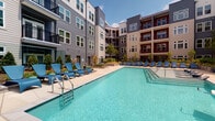 This amenity space is set within a contemporary multi-story building complex, centered around a striking courtyard design that creates a cohesive indoor-outdoor living environment. The architectural facade combines contrasting materials — gray and beige horizontal siding paired with bold red and neutral paneling — giving the complex a modern, dynamic appearance with large windows and private balconies overlooking the courtyard.

At the heart of the outdoor area is a generously sized resort-style swimming pool with crystal-clear blue water, accessible via built-in entry steps. The surrounding pool deck is finished in light-toned concrete pavers and is well-appointed with rows of blue lounge chairs, ideal for relaxing under open skies. Lush landscaping, including raised stone-edged planter beds with manicured shrubs and young trees, adds greenery and visual warmth throughout the courtyard.

Transitioning indoors, the covered lounge area seamlessly connects to the outdoor space through floor-to-ceiling glass sliding doors, allowing abundant natural light to flood the interior. The lounge features light wood-style flooring, shiplap-accented walls, and modern ceiling fans overhead. Comfortable seating arrangements with neutral sofas and vibrant accent pillows, alongside a wall-mounted television, create an inviting communal gathering space throughout the day.