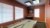 This single-floor recreational space is designed with entertainment and leisure in mind. The room's most striking feature is its bold terracotta-red walls, which lend a warm, energetic ambiance to the entire space. A full-size pool table takes center stage, complete with cues and a racked set of billiard balls, making it an immediate focal point for any visitor.

Overhead, two large flush-mounted grid lighting fixtures provide bright, even illumination throughout the room, ensuring excellent visibility for gameplay. Multiple windows dressed with white horizontal blinds line the walls, allowing natural light to filter in and complement the artificial lighting.

A decorative framed artwork depicting a lone figure in a dramatic spotlight scene adds a touch of artistic character to the space, reinforcing the room's entertainment theme. A small round table accompanied by stools is tucked neatly into one corner, offering a spot for casual seating or refreshments between games.

Dark carpet flooring grounds the room and contrasts effectively with the warm wall tones, while the overall layout is open and functional. This game room strikes a balance between purposeful design and recreational comfort, making it well-suited as a communal or private leisure area.