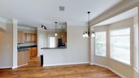 This single-floor home showcases a thoughtfully designed open-concept layout that effortlessly connects the kitchen, dining, and living areas into one cohesive and functional space. The kitchen features warm honey-toned wood cabinetry, dark countertops, and a raised breakfast bar peninsula that serves as both a prep area and a casual dining spot. Stainless steel appliances add a modern touch, while a dedicated track lighting system ensures the kitchen remains bright and well-lit. Crown molding runs throughout, lending an elegant and polished finish to every room. The dining area is anchored by a classic multi-arm chandelier and benefits from generous natural light streaming in through multiple windows dressed with white blinds. Hardwood flooring flows continuously across the space, adding warmth and visual continuity. The neutral, light beige wall tones create a calm and inviting ambiance that complements the natural wood tones throughout. Pendant lighting over the kitchen bar adds stylish accent illumination, while the open sightlines between spaces make the home feel expansive and airy. This home offers a clean, move-in-ready aesthetic with timeless architectural details.