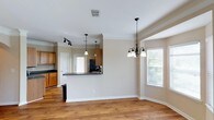 This single-floor home showcases a thoughtfully designed open-concept layout that effortlessly connects the kitchen, dining, and living areas into one cohesive and functional space. The kitchen features warm honey-toned wood cabinetry, dark countertops, and a raised breakfast bar peninsula that serves as both a prep area and a casual dining spot. Stainless steel appliances add a modern touch, while a dedicated track lighting system ensures the kitchen remains bright and well-lit. Crown molding runs throughout, lending an elegant and polished finish to every room. The dining area is anchored by a classic multi-arm chandelier and benefits from generous natural light streaming in through multiple windows dressed with white blinds. Hardwood flooring flows continuously across the space, adding warmth and visual continuity. The neutral, light beige wall tones create a calm and inviting ambiance that complements the natural wood tones throughout. Pendant lighting over the kitchen bar adds stylish accent illumination, while the open sightlines between spaces make the home feel expansive and airy. This home offers a clean, move-in-ready aesthetic with timeless architectural details.