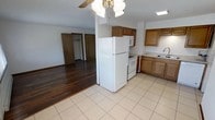 This single-floor residence offers a practical and functional layout designed for comfortable everyday living. The kitchen is equipped with warm-toned wood cabinetry, a full-size refrigerator, electric range, built-in microwave, and a sink with ample counter space. Light ceramic tile flooring runs throughout the kitchen, creating a clean and easy-to-maintain surface. The kitchen flows naturally into the adjacent living area, which features rich, dark hardwood flooring that adds warmth and character to the space. A ceiling fan with a light fixture is centrally positioned overhead, providing both air circulation and ambient lighting to the shared area. The open sightlines between the kitchen and living space create a sense of connectedness and ease of movement throughout the main living area. Beyond the living space, a hallway leads to additional rooms, with doorways visible that suggest access to a closet and additional living quarters. The overall layout is straightforward and well-organized, making efficient use of the available space while maintaining a welcoming and livable atmosphere throughout.