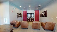 This media room is designed to deliver an immersive entertainment experience within a single-floor layout. The room centers around a large flat-screen television mounted on a media console, flanked by dramatic crimson curtain panels that evoke the ambiance of a classic cinema. Recessed lighting in the smooth ceiling provides adjustable illumination, complemented by crown molding that adds a polished architectural finish. The seating arrangement consists of multiple generously sized leather armchairs positioned in a semi-organized layout facing the screen, ensuring clear sightlines from every seat. Cinema-themed wall art on both sides of the room reinforces the theatrical aesthetic, featuring classic movie and theater imagery. The neutral-toned walls and patterned carpet flooring contribute to a warm, inviting atmosphere. The overall design balances comfort and functionality, making the space well-suited for group movie screenings or casual media viewing. The thoughtful combination of décor elements, from the stage-curtain draping to the curated artwork, creates a cohesive, purpose-driven environment that pays homage to traditional theater culture.