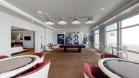 This well-appointed single-floor amenity space is thoughtfully divided into multiple distinct zones designed for socializing and entertainment. The main lounge area features a generously sized sectional sofa in cream upholstery, accented with vibrant jewel-toned throw pillows, patterned ottomans, and a dark rectangular coffee table. A ceiling fan with recessed lighting overhead and large windows with double doors provide ample natural light and direct access to an outdoor area. An adjacent kitchenette offers dark wood cabinetry, stainless steel appliances, and bar-height seating.

The billiards room is a striking highlight, centered around a full-size pool table with a red felt surface beneath a multi-pendant industrial-style chandelier. Bold black-and-white photographic artwork of classic Hollywood icons lines the walls, lending a sophisticated retro character. The card room features multiple round gaming tables surrounded by stylish white and burgundy upholstered chairs, with pendant lighting overhead and large windows offering views of the surrounding cityscape. Throughout, light gray walls, crown molding, and warm carpeting create a cohesive and inviting atmosphere.