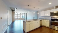 Located at 1059 Old River Rd in Cleveland, OH, this modern single-floor residence offers a thoughtfully designed open-concept layout with contemporary finishes throughout. The kitchen is a standout feature, equipped with stainless steel appliances including a range, microwave, and refrigerator, complemented by sleek white cabinetry, granite countertops, and rich hardwood flooring. A central island with a built-in sink and additional cabinet storage enhances both functionality and style, while pendant lighting adds a refined touch overhead. The expansive living area flows seamlessly from the kitchen, highlighted by an exposed concrete ceiling with industrial-style ductwork that adds an urban loft aesthetic. Large floor-to-ceiling windows flood the space with natural light and frame impressive views of the Cleveland skyline. The bedroom features plush carpeting, recessed lighting, a sliding glass door opening to a private balcony with city views, and direct access to a well-appointed bathroom. The bathroom includes a white vanity with granite countertops, a large mirror, tile flooring, and a soaking tub. A closet with built-in shelving provides additional storage. The overall ambiance blends industrial character with modern comfort in the heart of Cleveland.