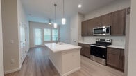 This single-floor residence is designed with a contemporary open-concept layout that seamlessly connects the kitchen and living areas. The kitchen serves as a central hub, anchored by a large white waterfall island with an undermount sink and pendant lighting overhead. Rich dark-brown shaker cabinetry lines the walls, complemented by white quartz countertops, a subway tile backsplash, and a full suite of stainless steel appliances, including a range, over-the-range microwave, and refrigerator. Light-toned wood-style flooring runs throughout the main living areas, creating a cohesive and clean aesthetic.

The living area extends naturally from the kitchen, featuring a ceiling fan and multiple windows dressed with blinds that allow ample natural light. A dedicated in-unit laundry nook with a stacked washer and dryer adds everyday convenience.

The bedroom is bright and well-proportioned, with dual windows providing outdoor views and generous natural light. It is served by a spacious walk-in closet outfitted with wire shelving and hanging rods for organized storage.

The bathroom features a dark-wood vanity with a white countertop, a large mirror, and a combination tub and shower with classic white subway tile surround, all finished with light flooring consistent with the rest of the unit.