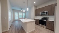 This single-floor residence is designed with a contemporary open-concept layout that seamlessly connects the kitchen and living areas. The kitchen serves as a central hub, anchored by a large white waterfall island with an undermount sink and pendant lighting overhead. Rich dark-brown shaker cabinetry lines the walls, complemented by white quartz countertops, a subway tile backsplash, and a full suite of stainless steel appliances, including a range, over-the-range microwave, and refrigerator. Light-toned wood-style flooring runs throughout the main living areas, creating a cohesive and clean aesthetic.

The living area extends naturally from the kitchen, featuring a ceiling fan and multiple windows dressed with blinds that allow ample natural light. A dedicated in-unit laundry nook with a stacked washer and dryer adds everyday convenience.

The bedroom is bright and well-proportioned, with dual windows providing outdoor views and generous natural light. It is served by a spacious walk-in closet outfitted with wire shelving and hanging rods for organized storage.

The bathroom features a dark-wood vanity with a white countertop, a large mirror, and a combination tub and shower with classic white subway tile surround, all finished with light flooring consistent with the rest of the unit.