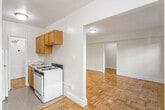 This single-floor residence is defined by its warm parquet wood flooring, which runs consistently throughout the living areas and bedroom, adding a timeless character to the space. The layout is open and functional, with a generously sized living area that benefits from natural light streaming through multiple windows fitted with wall-unit air conditioning and a classic radiator for year-round climate control. Neutral, cream-toned walls keep the interior feeling bright and airy.

An arched passageway connects the living area to the rest of the unit, providing a subtle architectural detail that adds charm. The galley-style kitchen is efficiently arranged, featuring warm wood cabinetry on both sides, a gas range, a full-size refrigerator, and a stainless steel sink with ample counter space. A door at the end of the kitchen leads to a dedicated storage or utility area. The flooring in the kitchen transitions to a speckled terrazzo-style surface, practical for the cooking space.

The bathroom is clean and straightforward, finished in white with a full tub-shower combination, toilet, and vanity sink. Terrazzo-style flooring continues in this space, complementing the bright, minimal aesthetic. Overall, the unit offers a cohesive, well-maintained interior with functional spaces throughout.
