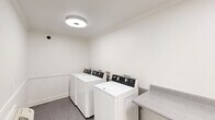 Located at 3320 Edenborn Ave in Metairie, Louisiana, this shared laundry facility offers a clean, efficiently organized space designed for practical, everyday use. The room is finished with crisp white walls accented by classic chair rail molding and crown molding at the ceiling, lending a polished, well-maintained appearance throughout.

The laundry room is equipped with multiple top-loading Maytag washing machines arranged in a row along one wall, providing ample capacity for residents. Opposite the washers, two sets of stacked dryer units offer convenient and space-efficient drying options. A built-in granite-style laminate folding counter runs along the wall, providing a dedicated surface for sorting and folding laundry.

The space is lit by flush-mount ceiling light fixtures that provide even, functional illumination. A wall-mounted air conditioning unit ensures comfortable conditions year-round. An exterior door with a glass panel allows for natural light and easy access, while a fire extinguisher mounted on the wall underscores the room's attention to safety. The neutral-toned flooring is durable and easy to maintain, complementing the room's clean and utilitarian aesthetic. Overall, this laundry facility is thoughtfully designed to serve residents with efficiency and comfort.