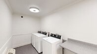Located at 3320 Edenborn Ave in Metairie, Louisiana, this shared laundry facility offers a clean, efficiently organized space designed for practical, everyday use. The room is finished with crisp white walls accented by classic chair rail molding and crown molding at the ceiling, lending a polished, well-maintained appearance throughout.

The laundry room is equipped with multiple top-loading Maytag washing machines arranged in a row along one wall, providing ample capacity for residents. Opposite the washers, two sets of stacked dryer units offer convenient and space-efficient drying options. A built-in granite-style laminate folding counter runs along the wall, providing a dedicated surface for sorting and folding laundry.

The space is lit by flush-mount ceiling light fixtures that provide even, functional illumination. A wall-mounted air conditioning unit ensures comfortable conditions year-round. An exterior door with a glass panel allows for natural light and easy access, while a fire extinguisher mounted on the wall underscores the room's attention to safety. The neutral-toned flooring is durable and easy to maintain, complementing the room's clean and utilitarian aesthetic. Overall, this laundry facility is thoughtfully designed to serve residents with efficiency and comfort.