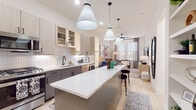 This thoughtfully designed single-floor residence showcases a seamless blend of modern aesthetics and functional living. The heart of the home is a striking open-concept kitchen, anchored by an expansive white quartz island with bar seating. Two-tone cabinetry pairs light wood upper cabinets with charcoal lower cabinets, complemented by a crisp subway tile backsplash and stainless steel appliances, including a gas range, dishwasher, built-in microwave, and a beverage cooler. Statement pendant lights hang above the island, adding a bold design touch. The kitchen flows effortlessly into the living area, where light hardwood flooring, neutral walls, and generous windows fill the space with natural light. A ceiling fan and wall-mounted TV enhance the comfort and functionality of the living room. The bedrooms offer a serene retreat, each with light hardwood floors, large windows with elegant draping curtains, and ample closet space. The bathroom is well-appointed with a dual-sink vanity featuring grey cabinetry, black hardware accents, large frameless mirrors, and a soaking tub surrounded by marble-look tile. Throughout the residence, clean lines and a warm, neutral palette create a cohesive and inviting atmosphere.