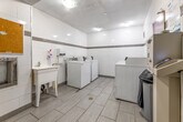 This shared laundry facility is a well-organized, single-room space designed to serve building residents efficiently. The room features white large-format ceramic wall tiles accented by a horizontal grey tile border that runs along the perimeter, lending a clean and orderly aesthetic throughout. The floor is finished with light grey porcelain tiles in a linear pattern, adding a modern touch to the utilitarian space.

The room houses multiple top-loading commercial washers and dryers arranged in two rows along opposing walls, each unit equipped with a card or coin-operated payment system managed through Sparkle Solutions. A dedicated payment kiosk is conveniently positioned within the room for easy access. A white utility sink with a faucet is mounted along one wall, accompanied by a wall-mounted soap dispenser and paper towel unit, supporting laundry prep needs.

Additional amenities include a centrally placed waste bin, a cork bulletin board for resident notices, and clearly posted service instructions and contact information for machine-related inquiries. Overhead flush-mounted lighting ensures the space is well-lit. Ventilation is provided through wall-mounted HVAC grilles. A standard door with a self-closing mechanism provides secure and easy entry and exit, making this laundry room a practical and functional shared amenity.