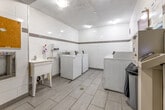 This shared laundry facility is a well-organized, single-room space designed to serve building residents efficiently. The room features white large-format ceramic wall tiles accented by a horizontal grey tile border that runs along the perimeter, lending a clean and orderly aesthetic throughout. The floor is finished with light grey porcelain tiles in a linear pattern, adding a modern touch to the utilitarian space.

The room houses multiple top-loading commercial washers and dryers arranged in two rows along opposing walls, each unit equipped with a card or coin-operated payment system managed through Sparkle Solutions. A dedicated payment kiosk is conveniently positioned within the room for easy access. A white utility sink with a faucet is mounted along one wall, accompanied by a wall-mounted soap dispenser and paper towel unit, supporting laundry prep needs.

Additional amenities include a centrally placed waste bin, a cork bulletin board for resident notices, and clearly posted service instructions and contact information for machine-related inquiries. Overhead flush-mounted lighting ensures the space is well-lit. Ventilation is provided through wall-mounted HVAC grilles. A standard door with a self-closing mechanism provides secure and easy entry and exit, making this laundry room a practical and functional shared amenity.