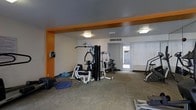 This fitness center offers a dedicated space for a comprehensive workout experience. The room is defined by its distinctive orange accent trim that frames the ceiling perimeter, adding a bold, energetic character to the otherwise neutral color palette of white walls and a gray ceiling. Overhead, flush-mounted square light fixtures provide consistent, bright illumination throughout the space.

The gym is equipped with a multi-station cable and weight machine as its centerpiece, supporting a wide range of strength training exercises. Complementing this is a stair climber or elliptical machine, a dumbbell rack, and additional free weights and exercise mats arranged along the floor, offering versatility for various fitness routines.

A glazed door with plantation shutters at the far end of the room allows natural light to filter in while maintaining privacy, and a ceiling-mounted HVAC vent ensures comfortable climate control during workouts. The carpeted flooring throughout provides a practical, cushioned surface suitable for floor exercises. The layout is open and functional, allowing users to move freely between equipment stations. Overall, the space delivers a practical and well-appointed environment for fitness and physical training.