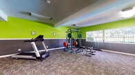 This fitness center offers a energetic and functional workout environment designed to accommodate a variety of exercise routines. The space is immediately distinguished by its bold two-tone wall treatment, featuring vivid lime green on the upper half and charcoal gray on the lower half, creating a visually dynamic and motivating atmosphere. Durable, patterned carpet tile flooring covers the entire workout area, providing comfort and traction underfoot.

The center is organized to support both strength training and cardiovascular exercise. A multi-function cable and Smith machine serves as the centerpiece of the strength training area, accompanied by a weight bench and a full rack of dumbbells, offering a comprehensive free-weight and resistance training setup. On the cardio side, a treadmill and an elliptical trainer provide options for aerobic workouts, along with a vertical knee raise station for core training.

Natural light filters in through multiple windows, offering views of the surrounding outdoor landscape, while overhead fluorescent fixtures ensure consistent interior lighting throughout the day. The textured ceiling adds an additional layer of architectural character to the space. The open, single-floor layout allows for ample room to move freely between equipment stations, making this fitness center a practical and inviting facility for daily exercise.