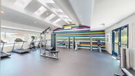 This fitness center in Escondido, California offers a well-equipped, single-floor exercise space designed to support a comprehensive workout experience. The open layout accommodates a wide variety of equipment, including Matrix treadmills, elliptical trainers, a stationary bike, cable and weight machines, and a dedicated functional training area stocked with medicine balls and resistance tools on a wall-mounted rack.

The interior design is a standout feature, anchored by a bold, multicolored horizontal striped accent wall in vivid shades of teal, pink, yellow, and white that energizes the space. Contrasting this, a separate alcove is finished with a deep teal accent wall, creating a distinct zone for functional fitness activities. The flooring throughout is durable rubber gym matting in a neutral speckled gray, ideal for high-traffic exercise use.

Architectural interest is provided by vaulted ceilings with exposed beam detailing and a series of skylights that flood the space with natural light. Recessed lighting supplements the brightness throughout. Large windows and glass double-entry doors along the perimeter offer views of the surrounding outdoor area, further enhancing the open, airy atmosphere. The overall ambiance is modern, vibrant, and well-maintained, making it a functional and visually stimulating environment for fitness enthusiasts.