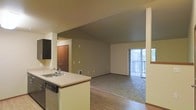 This single-floor space features an open-concept layout that seamlessly connects the kitchen and living areas. The kitchen is anchored by a peninsula with a light-toned countertop, an undermount sink with a standard faucet, and an integrated dishwasher. Dark upper cabinetry provides contrast against the neutral walls and lighter lower fixtures. The kitchen transitions naturally into the main living area, which is finished with plush carpeting, creating a distinct yet connected zone for relaxation and entertainment. Hardwood-style flooring is present in the kitchen and entry areas, adding warmth and visual variety to the space. A sliding glass door at the far end of the living area floods the room with natural light and provides access to what appears to be a private balcony or patio, offering outdoor views and ventilation. The entry door, finished in warm wood tones, is visible from the kitchen, contributing to the cohesive flow of the layout. Neutral wall tones throughout the space give it a clean, versatile aesthetic. The open floor plan and strategic placement of windows and doors contribute to a bright, airy ambiance across this well-organized single-level unit.
