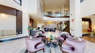Located in Franklin, Tennessee, this impressive community clubhouse and leasing facility delivers an upscale, hotel-inspired experience across a generously sized single-floor layout. The grand entrance lobby immediately commands attention with its soaring double-height ceilings, floor-to-ceiling windows that flood the space with natural light, and large-format marble-look porcelain tile flooring. A sweeping staircase with warm wood and metal railings leads to an open mezzanine level, framed by sleek glass balustrades overlooking the main floor below.

The reception area features a striking marble-topped counter accented by deep purple walls and geometric gold pendant lighting. Multiple distinct seating areas are thoughtfully arranged throughout, including a curved plum-toned sectional sofa set atop a bold zebra-print rug beneath a mid-century-modern sputnik chandelier, and a tufted charcoal armchair grouping centered around a linear fireplace with curated artwork above. A curved built-in sofa nook with warm wood ceiling detail and gold accent tables provides additional lounge space.

The leasing office area transitions to rich hardwood-style flooring, exposed beam ceilings, and bar-height seating along a long dark reception counter. Throughout, the design blends contemporary elegance with warm, inviting textures, creating a refined and welcoming environment.