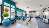 Located at 15645 N 35th Ave in Phoenix, AZ, this fitness center offers a comprehensive, single-floor workout facility designed to accommodate a wide range of exercise routines. The space is immediately distinguished by its bold teal accent walls, which contrast with light gray and white finishes to create an energizing yet clean atmosphere. Large windows along multiple walls flood the interior with natural light and provide views of the outdoor pool area, accessible through a set of elegant double glass doors framed by a arched transom window.

The cardio zone features multiple treadmills, elliptical trainers, and a rowing machine, all arranged to face mounted flat-screen televisions for an engaging workout experience. A dedicated strength training area is equipped with a multi-functional cable machine and a weight bench, while a well-stocked dumbbell rack—reflected in full-length mirrors—provides options for free weight exercises. Resistance bands, medicine balls, stability accessories, and yoga mats are neatly organized on wall-mounted racks, supporting functional and flexibility training.

The flooring transitions between patterned carpet tiles and hard surface areas, delineating different workout zones effectively. Recessed lighting complements the natural light throughout. The overall design is modern, practical, and inviting, making this fitness center a fully functional amenity for residents in the Phoenix community.