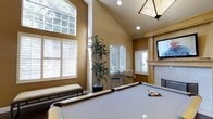 This inviting game room offers a thoughtfully designed recreational space highlighted by dramatic vaulted ceilings that lend an airy, open feel to the interior. A full-size billiards table takes center stage, positioned beneath a stylish pendant light fixture and recessed lighting that ensures excellent illumination for gameplay. The warm, earthy-toned walls create a welcoming ambiance throughout the room.

Multiple windows fitted with classic white plantation shutters line the walls, allowing an abundance of natural light to filter in while offering privacy and light control. The soaring ceiling height is further accentuated by a tall bank of upper windows, framing views of the surrounding greenery outside.

A cozy fireplace with a light wood mantel anchors one wall, accompanied by a wall-mounted flat-screen television, creating an ideal setting for relaxed entertainment. A high-top bistro table with modern bar stools provides additional seating and a casual gathering spot. A decorative potted plant adds a natural, refreshing element, while a cushioned bench along one wall offers comfortable additional seating. The overall design balances recreational functionality with a warm, inviting aesthetic.