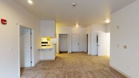 This single-floor unit is thoughtfully laid out to maximize functionality across its key living spaces. The open-concept living area serves as the heart of the unit, featuring wall-to-wall neutral carpeting, white walls, and a seamlessly connected kitchen that opens via a peninsula counter with an overhang suitable for casual seating. The galley-style kitchen is efficiently arranged with light wood cabinetry lining both walls, white laminate countertops, a stainless steel sink, and appliances including an electric range and refrigerator. Vinyl tile flooring completes the kitchen's clean, practical aesthetic.

Two carpeted bedrooms each benefit from natural light provided by windows overlooking the outdoors, wall-unit air conditioners, and baseboard heating. Notably, one bedroom features dual reach-in closets, offering generous storage. Both rooms maintain a neutral, bright palette.

The unit includes two full bathrooms, each featuring a tub-shower combination with white square tile surrounds, chrome fixtures, and grab bars for added accessibility. One bathroom includes a wood-toned vanity with countertop. Throughout, the unit presents a clean, functional design with a consistent neutral color palette and practical finishes.