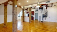 Located at 1329 Jefferson St in Lynchburg, VA, this two-story loft unit offers a compelling blend of historic industrial character and contemporary updates. The main floor is generously proportioned, anchored by striking exposed timber posts and beams that speak to the building's original construction. Warm hardwood floors run throughout, complementing the open-concept kitchen and living area. The kitchen is well-appointed with rich cherry cabinetry, granite countertops, stainless steel appliances including an electric range and built-in microwave, and a peninsula with a live-edge wood countertop detail. A dramatic spiral staircase connects the main floor to the upper level, where two loft spaces provide flexible use — one featuring additional exposed timber columns and the other showcasing a whitewashed brick accent wall. The bedroom is tucked under a sloped roofline with exposed beams and a large window that brings in natural light. The bathroom is clean and functional, finished with white tile, a tub-shower combo, and matching cherry vanity. Ceiling fans are installed throughout, and the angled rooflines give each room a distinctive, characterful feel unique to this historic Lynchburg property.